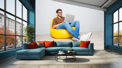 Happy young man sitting in yellow pouf  and using laptop Wall mural