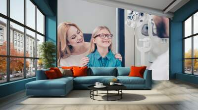 Healthcare, people, eyesight and vision concept. Small cute blond girl is trying on glasses, mom and doc are smiling, she is happy Wall mural