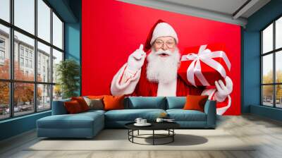 Ho ho! Holly jolly x mas! Noel magic! Funny kind astonished saint nicholas in traditional head wear is presenting a wrapped gift with white tape bow with forefinger up, isolated on red background Wall mural