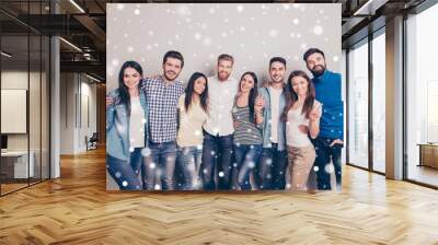 In winter eight cheerful young people embracing and smiling in casual clothes. Ladies are showing two fingers sign, snowflakes background Wall mural