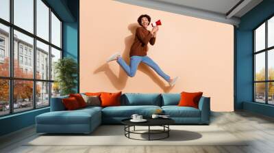 Joyful young man jumps with excitement holding a megaphone against a beige background in casual attire perfect for promotional concepts. Wall mural