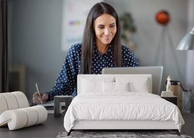 Photo of attractive cheerful young business woman write notebook look computer sit desk chief leader indoors in office Wall mural