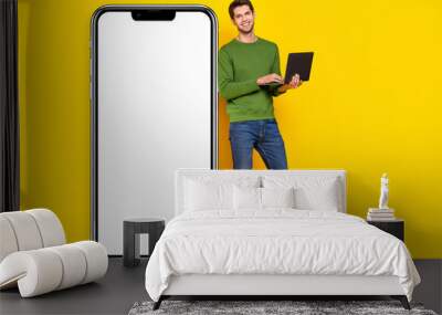 Photo of attractive cute young guy wear green sweater working modern device empty space isolated yellow color background Wall mural