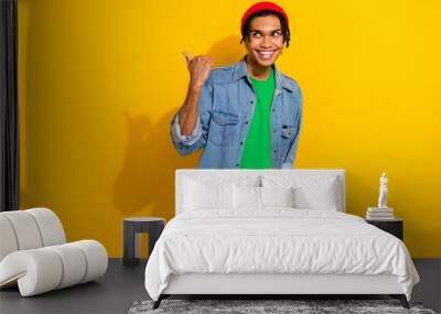 Photo of attractive dreamy man wear jeans shirt red hat looking pointing thumb empty space isolated yellow color background Wall mural