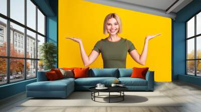 Photo of cheerful girl with bob hair dressed khaki t-shirt palms comparing products empty space isolated on yellow color background Wall mural