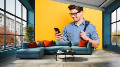 Photo of cheerful handsome attractive man browsing through telephone smiling toothily with backpack behind isolated vivid color background in jeans denim Wall mural