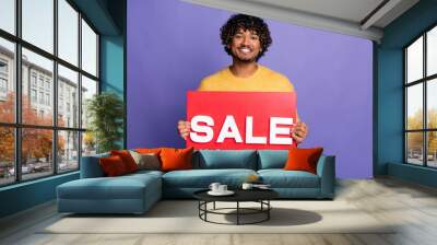 Photo of cheerful latin man wear stylish yellow clothes arms hold red placard sale news isolated on purple color background Wall mural