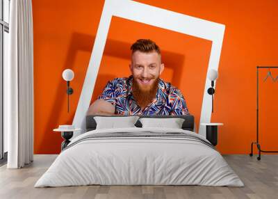 Photo of cool cheerful guy with red beard wear print shirt posing in white frame hold arms crossed isolated on orange color background Wall mural