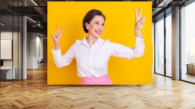 Photo of cute funny age woman dressed white shirt smiling showing v-sign dancing looking empty space isolated yellow color background Wall mural