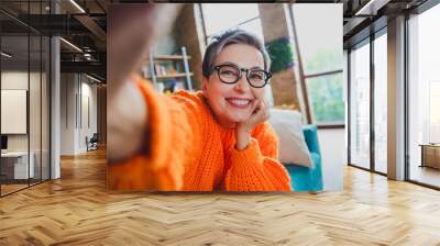 Photo of dreamy pretty elderly lady wear orange pullover spectacles arm hand cheek recording video indoors apartment room Wall mural