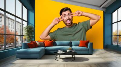 Photo of excited carefree man wear khaki t-shirt showing two v-signs cover eyes isolated yellow color background Wall mural