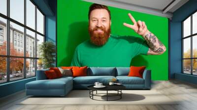 Photo of excited cheerful man beaming smile demonstrate v-sign isolated on green color background Wall mural