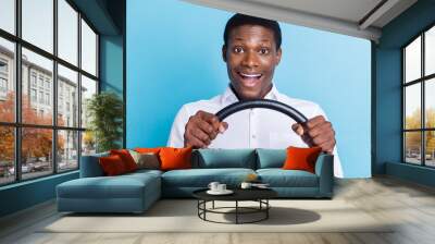 Photo of funny impressed dark skin man wear white shirt smiling holding steering wheel isolated blue color background Wall mural