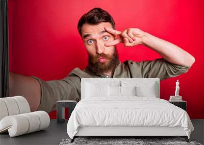 Photo of handsome attractive man wearing grey shirt showing you v-sign near eye as he plumps you while isolated with red background Wall mural