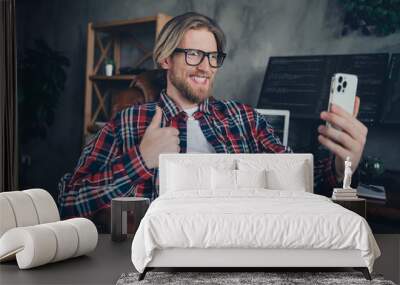 Photo of happy blond hair young programmer guy sitting on armchair at office with smartphone showing thumb up photographin himself Wall mural