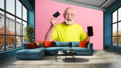 Photo of old excited cheerful smiling man wear lime t-shirt hold smartphone credit card isolated on pink color background Wall mural