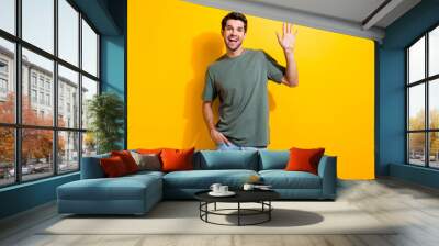 Photo of optimistic man with bristle dressed khaki t-shirt waving palm say hi hand in pocket isolated on vibrant yellow color background Wall mural