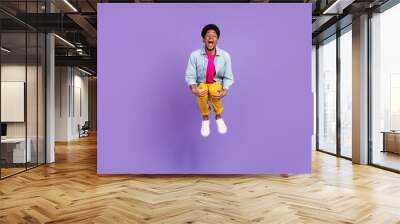 Photo of positive cheerful guy jump arms cuddle knees wear blue shirt pants shoes isolated purple color background Wall mural