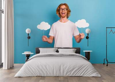 Photo of positive cheerful man hold two cloud paper card stick toothy smile isolated on blue color background Wall mural