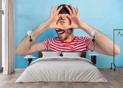 Photo of positive pretty young man dressed striped t-shirt showing fingers heart cover face isolated blue color background Wall mural