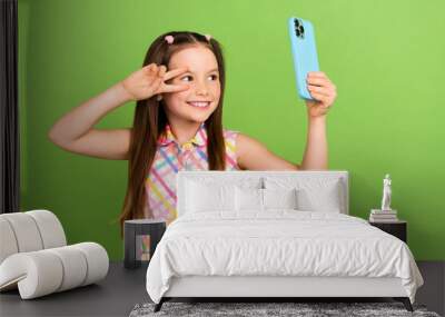 Photo of small charming schoolgirl hold smart phone take selfie demonstrate v-sign near eye isolated on green color background Wall mural