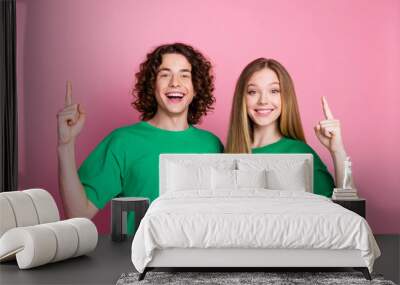 Photo of smiling happy curly haired teenage guy with girlfriend wear green t shirt direct fingers up isolated on pink color background Wall mural