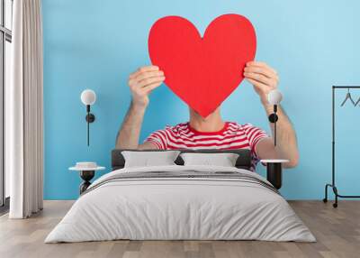 Photo of sweet shy young man dressed striped t-shirt red paper heart cover face head empty space isolated blue color background Wall mural