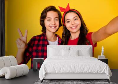 Photo of two small kids girl take selfie boy fingers show v-sign beaming smile isolated on yellow color background Wall mural