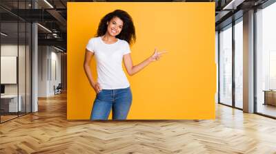 Photo of wavy cheerful girlfriend wearing white t-shirt pointing at empty space in jeans denim isolated vibrant yellow color background Wall mural