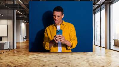 Photo of young dissatisfied grimace guy wearing yellow shirt holding mobile phone social media isolated on dark blue color background Wall mural