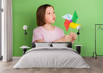 Photo of young little girl blow windmill pinwheel rotate toy isolated over green color background Wall mural