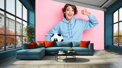 Photo of young nice pretty cute woman wear blue hoodie hold new football ball recommend showing finger thumb up like excited isolated on pink color background Wall mural