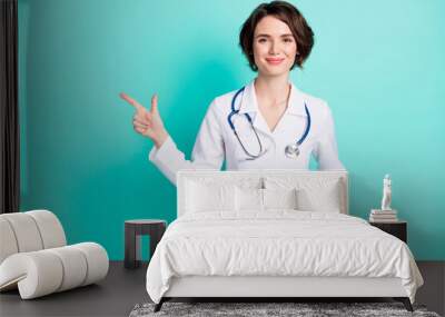 Photo of young woman doctor happy smile indicate finger empty space advert recommend select isolated over teal color background Wall mural