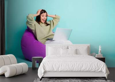 Photo portrait full body view of amazed screaming woman with laptop holding head with two hands sitting in purple chair isolated on vivid cyan colored background Wall mural