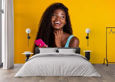 Photo portrait of african american woman with long wavy hair holding pink smartphone in two hands laughing wearing blue singlet isolated on vivid yellow colored background Wall mural