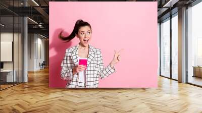 Photo portrait of excited woman pointing finger at blank space holding phone in one hand isolated on pastel pink colored background Wall mural