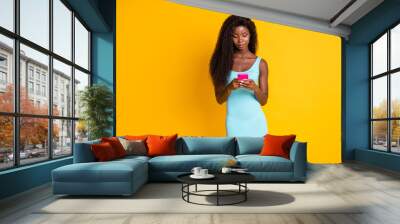 Photo portrait of focused african american woman holding pink phone in two hands browsing news wearing short blue dress isolated on vivid yellow colored background Wall mural