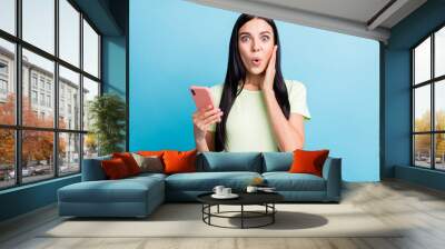 Photo portrait of impressed woman touching face cheek holding phone in one hand isolated on pastel blue colored background Wall mural