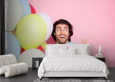 Photo portrait of man pointing finger looking at blank space holding balloons isolated on pastel pink colored background Wall mural