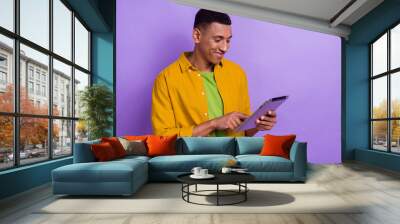 Photo portrait of nice young guy holding tablet gadget typing scrolling wear trendy yellow garment isolated on purple color background Wall mural