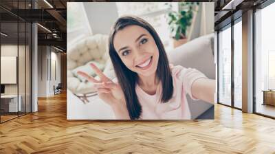 Photo portrait of overjoyed girl taking selfie showing v-sign with two fingers sitting on sofa indoors Wall mural