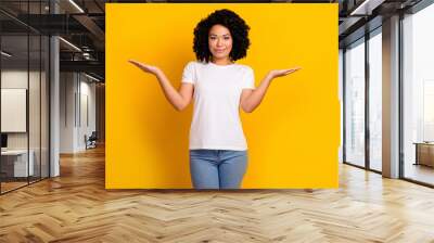 Portrait of adorable girl with curly hairdo wear white t-shirt arms comparing objects empty space isolated on yellow color background Wall mural