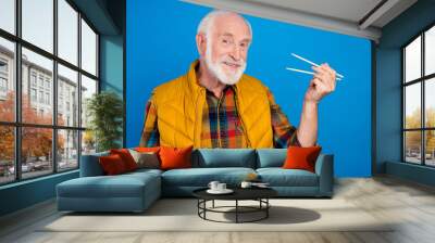 Portrait of attractive cheerful grey-haired man eating eastern food using sticks isolated over bright blue color background Wall mural