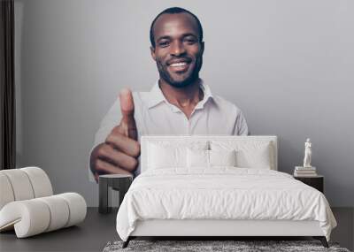 Portrait of cheerful, positive, handsome man with black skin, beaming smile in white shirt showing thumb up with finger to the camera isolated on grey background Wall mural