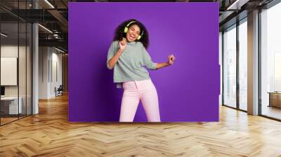 Portrait of cheerful afro american girl listen loud music use green headset enjoy sing song dance feel rejoice wear striped shirt pink pants trousers isolated over purple color background Wall mural