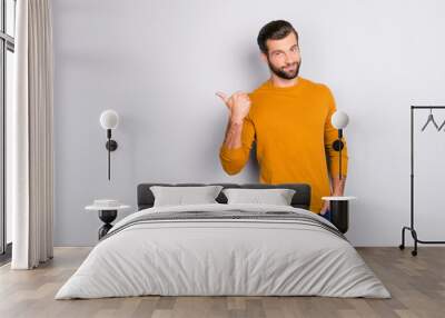 Portrait of cheerful attractive joyful confident guy keeping hand in pocket of jeans hinting pointing on copyspace behind his back isolated on gray background Wall mural