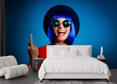 Portrait of cheerful provocative girl in headwear eyewear gesturing fuck off sign with middle finger isolated on blue background. Conflict insult dispute concept Wall mural