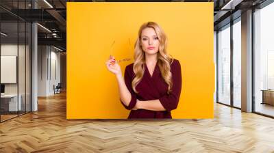 Portrait of experienced thoughtful lady in formal wear, holding glasses in hands, looking at camera isolated on yellow background Wall mural