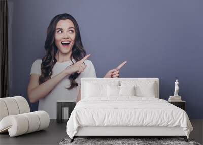 Portrait of magnificent lovely adorable stylish cheerful delighted curly-haired girl in casual white t-shirt, pointing aside up with two hands, opened mouth, copy-space, isolated over grey background Wall mural