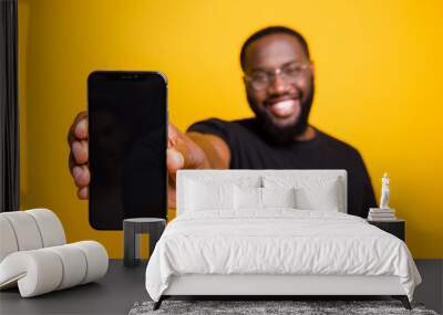 Portrait of positive afro american guy hold smartphone show modern technology gadget advertise promotion wear casual style outfit isolated over yellow color background Wall mural